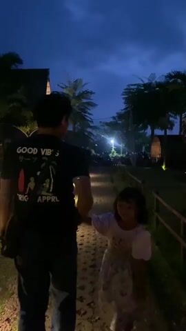 In a low-lit outdoor scene, a man and a young girl are walking hand-in-hand on a stone pathway, away from the camera. The man is wearing a black t-shirt with white text and a graphic on the back, and dark jeans. A camera bag is slung over his shoulder. The girl is wearing a dress and white sneakers. The sky is a deep blue, suggesting it is dusk or early evening. Palm trees and other tropical vegetation are visible along the path, as well as small buildings with thatched roofs. String lights and lanterns illuminate the path and surrounding area, creating a warm glow. Other people are walking further down the path, some obscured by the darkness.