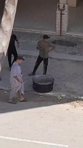 Several people are gathered around a large pot. An adult male wearing a light-colored robe and a cap is bending over the pot, appearing to be stirring or adding something to it with his right hand. To his left, a young boy wearing a blue shirt and shorts holds a smaller metal pot. Behind him, another adult male in a black t-shirt and dark pants gestures with his right hand, looking towards the pot. To the right of the adult in the robe, a man in a green t-shirt and a black cap stands holding a long tool, possibly a shovel or a pole, with his right hand on his hip. In the background, a building with brickwork is visible, along with what appears to be a manhole cover or an opening in the ground. The ground surface is rough and appears to be a mixture of dirt and pavement. The overall scene suggests an outdoor communal activity involving the preparation of food or a similar task.