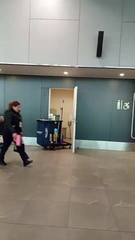 The video shows a hallway with tiled flooring and gray walls. On the left, a cleaning cart is visible with mops, buckets, and cleaning supplies. A person in a dark jacket and pants walks from left to right, carrying a pink bag. They pass by the cleaning cart and approach a doorway. The doorway leads to a room with a toilet and another cleaning cart. The video continues to pan across the hallway, showing another cleaning cart on the right. Signs indicating restrooms are visible on the wall.