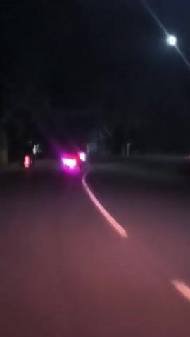The video captures a nighttime scene from a moving vehicle on an asphalt road in Gampongrayadagang, Indonesia. The road features a visible white dividing line that curves to the right. On the left side of the road, a vehicle with prominent bright pink or purple taillights is seen, followed by another vehicle with standard red taillights further ahead. As the video progresses, multiple sets of bright white headlights from oncoming vehicles become visible, creating strong lens flare effects that partially obscure the view. The overall scene is dark, typical of night driving, with vehicle lights serving as the primary illumination. No people or visible text are discernible. The weather appears clear.