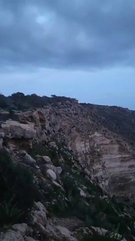 The video shows a cliffside overlooking a body of water. The cliff is layered with exposed rock formations and sparse green vegetation. The water appears calm and dark blue, stretching out to the horizon under an overcast sky. The sky is a mix of blue and gray clouds. The camera pans slowly to the right, revealing more of the coastline and the open water.