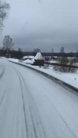 The video shows a snowy road on an overcast day. A small wooden structure with a green-topped spire stands to the side of the road, covered in snow. A snow-covered picnic table is next to it, also laden with snow. Bare trees and shrubs line the horizon, and a river or stream is visible behind the structure. Tire tracks are visible in the snow on the road. A road sign is visible on the left side of the road. The video appears to be taken from a moving vehicle.