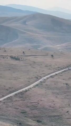 A white car drives on a dirt road through a vast, dry, grassy landscape. The terrain is characterized by rolling hills and distant mountains, all appearing arid and sparsely vegetated. The sky is overcast with a light, hazy tone. The camera pans slightly, revealing the emptiness and scale of the landscape.