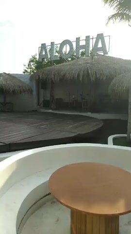 The video showcases an outdoor pool area with a tropical theme.  The pool water is a pale green color. White curved structures with built-in seating surround the pool.  Wooden tables are integrated into these seating areas. The area features wooden decks under thatched umbrellas, providing shaded seating. Palm trees are strategically placed, enhancing the tropical ambiance. In the background, a sign reading "ALOHA" is visible above a thatched-roof structure, suggesting a bar or stage area.  The video pans across the scene, highlighting the different elements of the pool area. The sky is partially cloudy.