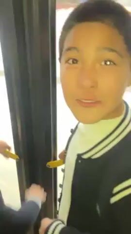 A person is standing in the doorway of what appears to be a bus. The person is young, has short hair, and is wearing a black jacket with white stripes. They look at the camera, smile, and then look out the window. They are holding onto a yellow handrail. Through the window, you can see a street with cars and other buildings.