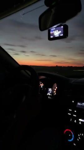 The video shows the interior of a car driving at dusk. The camera is positioned inside the car, looking forward through the windshield. The dashboard and steering wheel are visible in the foreground, partially illuminated by the car's interior lights. In the center of the windshield, a small screen, likely a GPS or dash cam, displays the speed as 141 mph. Outside the windshield, the sky is a gradient of deep blue and orange, indicating sunset or sunrise. A distant horizon line is visible, with faint lights suggesting a populated area. The road ahead appears to be a highway. The overall impression is of a high-speed drive during twilight.