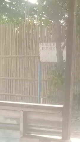 The video shows a close-up of a sign that reads "BENGE LISTRIK". The sign is white with black lettering. Behind the sign is a bamboo fence. There are trees and foliage visible. A wooden beam is in the foreground. The lighting suggests it is daytime.