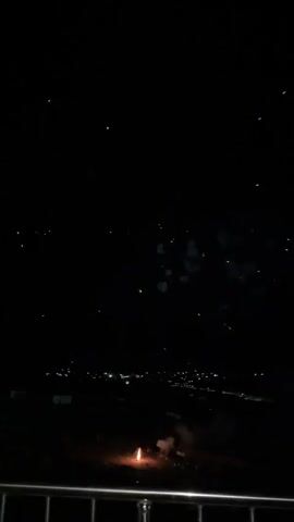 The video captures a nighttime fireworks display. The scene is set against a dark, clear sky, suggesting fair weather. In the foreground, a metal railing is visible, indicating the vantage point from which the recording was made. Below the railing, a dimly lit landscape unfolds with scattered lights suggesting a town or settlement in the distance, consistent with the Kaji-Say, Kyrgyzstan location.

The main subject is the fireworks. The display begins with a single rocket ascending and exploding into a vibrant burst of light. The initial rocket ignites and streaks upwards, leaving a trail of sparks. It then detonates, creating a large, radial burst of white and yellow fireworks, with some red accents scattered throughout. This is followed by a brief period of darkness, then another rocket launches and explodes similarly, though this second burst appears to have slightly more red and orange hues. The fireworks are the central focus, dominating the frame with their brilliant illumination against the dark backdrop.

The activity is clearly a fireworks show, likely a celebration or special event. The time of day is night, as evidenced by the darkness and the prominence of the artificial lights from the fireworks and the distant settlement. No people are visible in the frame, and the focus is entirely on the aerial pyrotechnics. The emotion evoked is one of excitement and visual spectacle, typical of fireworks displays. There is no visible text in the media.