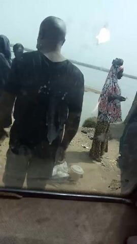 From inside a vehicle, the video shows several people by a lake.  A bald man in a black shirt and gray shorts gestures with his arms raised. There are children standing near him. A woman wearing a patterned headscarf and dress stands to the right. There are buckets and plastic bags on the ground, and a makeshift structure beside the woman. The lake is visible in the background. The windshield of the vehicle obstructs the view slightly.