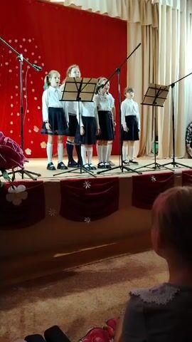 Certainly! Here's a concise description of the scene:

Five young girls stand on a small stage, singing into microphones. They are dressed in matching uniforms: white blouses, dark skirts, and white socks. Sheet music stands are placed in front of them. The stage is decorated with burgundy fabric and white snowflake accents. Behind them, there's a red curtain and white drapes. In the foreground, a girl with blonde hair is watching them.