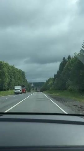 The video shows a view from inside a car driving on a two-lane road surrounded by trees under a cloudy sky. On the left side of the road, there are two trucks, one white and one red. On the right side, there is a dense line of evergreen trees. In the distance, more trees and a rolling landscape are visible, with two more trucks on the road. The sky is overcast with dark gray clouds.