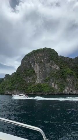 A speed boat is moving across dark blue water with a large, green, rocky island in the background. The sky is cloudy and gray. The speed boat is white with red and black accents and is leaving a white wake behind it. There are people visible on the speed boat. The camera is angled downwards from another boat.