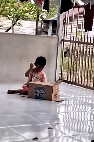 A young boy sits on a tiled floor next to an open cardboard box. He is wearing a pink and white t-shirt and red shorts. He holds a green object in his right hand and appears to be interacting with the box. The box has the word "AQUA" printed on it. The background shows a concrete wall, a metal fence, trees, and laundry hanging from a clothesline. The lighting suggests it is daytime.
