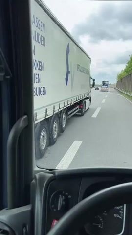 The video shows a view from inside the cab of a truck driving on a highway. Two other trucks are visible ahead, one is a tanker truck and the other is a standard semi-trailer. The sky is overcast and the road is divided into multiple lanes. A noise barrier is visible on the right side of the highway. The truck is moving at a moderate speed. The truck's dashboard shows the mileage, time, and outside temperature. There are no visible passengers. The camera is static and the focus is on the road ahead.