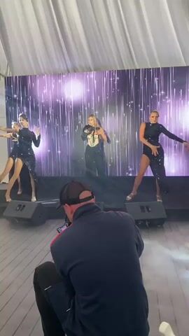 A video shows a female singer performing on stage with several dancers. The singer is in the center, wearing a sparkly corset and black pants, holding a microphone. To her left and right are two dancers in matching black sequined dresses with high slits. Two more dancers are visible further to the sides, also in similar outfits. They are all performing in front of a backdrop with a sparkling, starry, purple light effect. In the foreground, a person is crouched down, filming the performance with a phone. The stage is set up with black speakers. The environment appears to be indoors, possibly a venue with draped white curtains in the background.