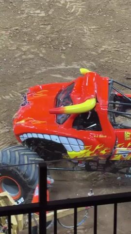 A monster truck with a red and yellow bull-like design, named "El Toro Loco," is parked on a dirt surface. A person in an orange jumpsuit is standing near the front tire of the truck. The truck has large tires and a cage structure in the back. The background appears to be an arena or exhibition space.