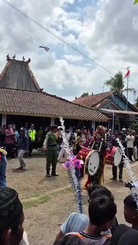 A street parade is underway, featuring a drum corps with young performers. The individuals in the foreground are wearing decorative headdresses and colorful costumes, some carrying drums. The crowd appears to be watching the parade from both sides of the street, with some individuals in uniform visible among them. The background shows traditional Indonesian houses with red tile roofs. The sky is partly cloudy, and there is a flag visible on a pole. The overall atmosphere seems festive.