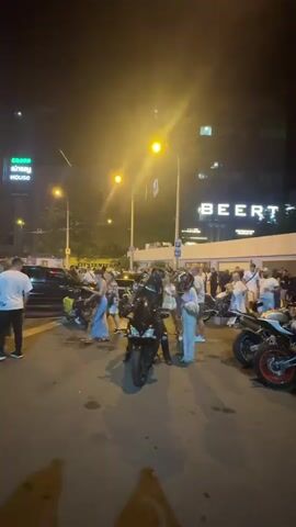 A nighttime street scene outside a building with a large illuminated sign reading "BEERT". Multiple people are gathered on the street, some on motorcycles. A person in a black motorcycle suit is sitting on a black motorcycle in the center of the frame. To the right, two people are on a white motorcycle with red accents on the wheels, and another person is standing next to them. Several other individuals are standing or walking around the motorcycles and near the building entrance. The environment appears to be an urban setting with streetlights and car headlights visible.