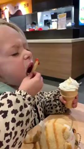 A young, fair-skinned girl with blonde hair, wearing a leopard-print jacket over a turquoise hoodie, is sitting at a wooden table in a fast food restaurant. She's eating a french fry dipped in ketchup while holding an ice cream cone in her other hand. A plush toy cat rests on the table in front of her. The restaurant has yellow and black signage, and a service counter with employees visible in the background.
