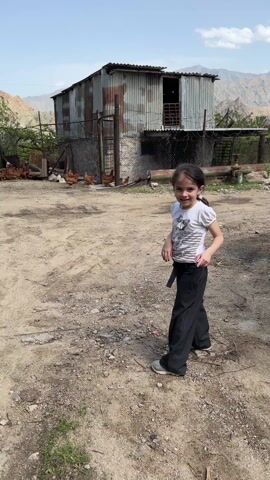 A young girl stands in a dirt yard and then runs towards a flock of chickens.  The girl is wearing a white t-shirt, black pants, and grey shoes, and has a white flower in her hair. She turns and smiles at the camera before running toward the chickens. The chickens are grouped together in front of a rusty metal shed surrounded by a chain link fence. Mountains are visible in the background.