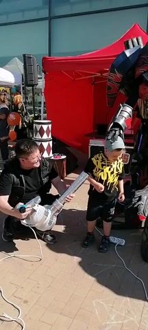 A man in a black shirt and vest kneels next to a child in a yellow and black shirt, holding a large silver toy gun. A large robot costume stands nearby. The robot appears to be Optimus Prime, and the child is laughing. The man wears glasses and has short dark hair. The child has short blonde hair. The robot is tall and dark, with red and blue accents and glowing lights. In the next shot, a robot costume, resembling Bumblebee, stands alone. The Bumblebee costume is yellow and black with silver accents. The background features a building and a red tent.