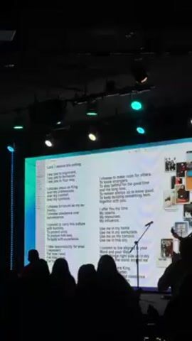 The scene is set in a dimly lit auditorium with a large screen displaying text. On the left side of the screen is a list titled "Lord, I receive this calling," with several lines of text underneath. On the right side are corresponding text blocks next to a collage of various photos. A microphone stands on a stand in front of the screen. Seated in the foreground are several silhouetted figures of people listening. Above, there are multiple stage lights, with some emitting blue and green colors. The overall atmosphere is one of a formal gathering or event.