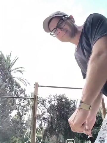 A man is seen doing pull-ups outdoors. He is wearing a black t-shirt, a beige baseball cap, glasses, and patterned shorts. He has a watch on his left wrist. The background features trees and a cloudy sky. The man is on a pull-up bar, and his body is captured in various stages of the exercise.