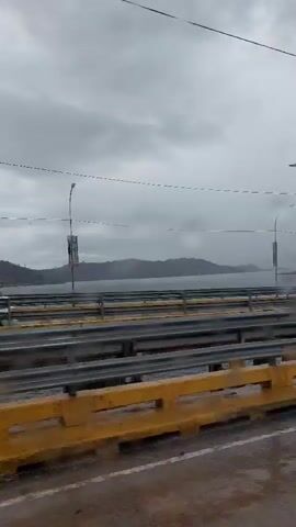 The video captures a view from inside a moving vehicle traveling along a multi-lane bridge under an overcast sky. The road surface is visibly wet, indicating rain or recent precipitation. Yellow concrete or plastic barriers are present on the side closest to the camera, followed by metal guardrails separating the lanes.

Tall utility poles with streetlights and overhead wires extend along the bridge. One rectangular sign on a pole is clearly visible, bearing the text "ISLA LARGA." In the background, a large body of water, likely Lake Valencia, spans out towards distant mountains or hills, which are partially obscured by clouds or haze.

During the clip, a white truck and subsequently a red semi-trailer truck pass by in the opposite direction on the bridge. The scene is consistent with a daytime view, despite the cloudy weather, in the vicinity of La Cabrera, Venezuela, near Isla Larga. Background audio features Latin music.