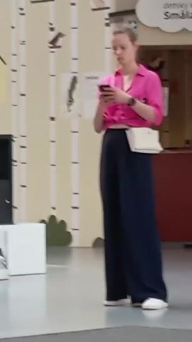 A young woman with blonde hair pulled back stands facing forward, looking down at her phone. She is wearing a bright pink cropped shirt and dark blue wide-leg pants. A white crossbody bag is slung across her right shoulder. She is in an indoor environment with a patterned wall behind her featuring images of birds and trees. A white sign with text in a foreign language is visible above her. To the right of the frame, another woman is partially visible, seated at a counter. The floor appears to be a light-colored tiled surface.