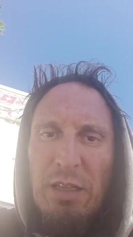 A close-up shot shows a man's face. He has dark hair, with some strands sticking up. He is wearing a grey hooded sweatshirt. He has a light beard and a missing tooth. The background is a bright blue sky and a white building with a red sign. The man is talking to the camera.