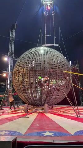 Inside a circus tent, the "Globe of Death" act features a large, spherical cage on wheels, secured by metal bars. Two motorcyclists ride inside the cage simultaneously, their bodies visible through the gaps in the latticework. A man in silver pants stands by the cage. The circus ring is decorated with red, white, and blue starburst patterns.  A spotlight shines from above, and a series of gymnastics rings hang near the top of the tent.
