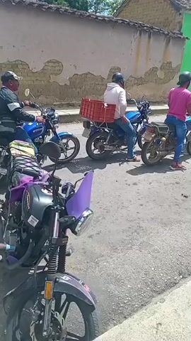 Several motorcycles are parked on a paved street, some with riders on them. In the background, a white pickup truck is also parked. People are visible around the vehicles. The area appears to be a town with buildings lining the street. A cell tower is visible in the distance. The weather is sunny with some clouds.