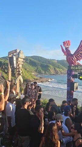 This video shows a crowd of people at a beach party during the daytime. There's a DJ booth decorated with vibrant purple and pink elements, topped with large, red, bird-like sculptures.  The DJs are visible behind their equipment, one in a white shirt and the other in a dark shirt. The audience is diverse, and many are dancing and looking towards the DJ. The location is a beach with green hills in the background and a clear blue sky. The ocean is visible, with waves gently rolling in. The atmosphere appears lively and energetic.