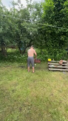 The video captures a man standing behind a barbecue grill in an outdoor, grassy setting with abundant greenery. He is shirtless, wearing grey three-quarter length pants and flip-flops. A large tattoo is visible on his upper back and left arm. The man appears to be tending to the coals in the grill with a tool.

The scene is set in a yard, possibly a backyard or a rural area, suggested by the trees and foliage. There is a wooden structure to the right of the grill with a yellow bowl and a bottle on it. The weather appears to be fair and the time of day is likely daytime, given the lighting.

The activity is focused on preparing the barbecue, with the man actively engaged with the grill. The overall impression is one of casual outdoor cooking. There is some visible text on the grill, partially obscured, but "K HOC" can be discerned.