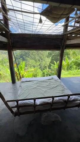 The video shows a bed with white sheets and a light green blanket. The bed appears to be suspended, possibly a hammock bed. In the background, there is a lush green jungle landscape with trees and vegetation. The sky is visible with some clouds. The structure above the bed appears to be made of bamboo and netting, suggesting an open-air or semi-open-air setting. The camera is stationary, and the view is from slightly above the bed, looking out towards the jungle.