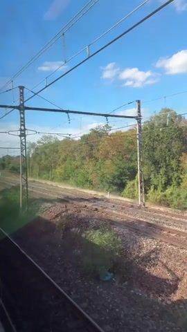 The video is filmed from a moving train, looking out the window to the right. The sky is blue with scattered white clouds. Overhead, train power lines are visible, supported by tall metal poles. In the foreground, there are train tracks and gravel. Beyond the tracks, there is a line of trees with autumn foliage, displaying shades of green, yellow, and orange. A white, rectangular structure is visible in the distance. As the video progresses, another train passes by on a parallel track. This passing train is white and blue and is in motion, making it appear blurred.