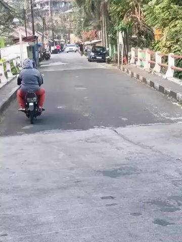 A motorcycle rider is seen from behind, heading up a paved road. A black pickup truck is also on the road, in front of the motorcycle. The road is lined with a bridge railing and trees. There are other vehicles and buildings visible in the background. The video appears to be taken from another vehicle, possibly a car, also moving on the road. The license plate of the pickup truck is visible, reading F 8285 AZ. The time stamps indicate the video is very short, likely just a few seconds long.