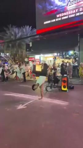 A young male breakdancer is performing on a street at night. He is wearing a white tank top and black pants. He is balancing on one hand and spinning his body in a circle. There are people watching him, and a speaker playing music nearby. The street is marked with a white arrow, and there are shops and palm trees in the background. The area is well-lit, with streetlights and the bright lights from the shops and a big digital display sign.