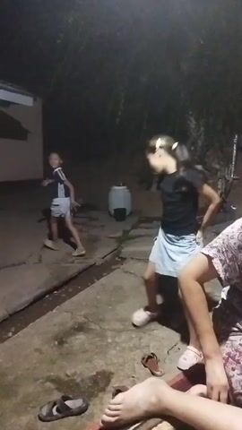 The video shows an outdoor scene at night. There are two young females in the foreground, one in a black t-shirt and light blue shorts, and another in a navy blue t-shirt and light blue shorts. The girl in the black t-shirt is facing away from the camera, dancing or playing, with her arms outstretched. The other girl is facing left, walking away from the camera. In the background, a light-colored building is visible, with a window on the left side. There are also some white pots on a ledge in the background, and a large white cylindrical object to the right of the dancing girl. The ground is paved and appears wet in places, with a drainage ditch running along the left side of the paved area. The scene is dimly lit, suggesting it is nighttime. Some sandals are visible on the ground in the foreground.