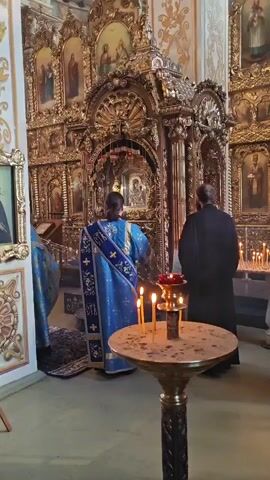 The video shows the interior of a church. Two figures in religious vestments stand facing an ornate altar. The figure in the foreground is wearing a bright blue robe with gold trim, and a gold sash. The figure behind them is wearing a dark robe. There are lit candles on a stand in the foreground, and more candles on the altar. The altar is decorated with gold trim and icons. The walls of the church are painted with religious imagery.