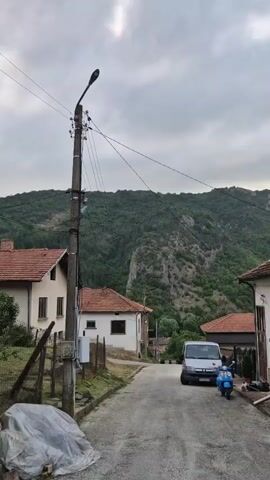 The video shows a quiet village street with houses on either side and a lush green mountain in the background. A white van and a blue scooter are parked on the side of the road. A utility pole with power lines stands on the left side of the street. The sky is cloudy, and the overall atmosphere is serene and peaceful.
