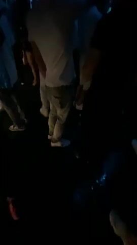 The video shows a group of people in a dimly lit nightclub. Blue neon lights are visible above the crowd, and there are also lights and speakers suspended from the ceiling. The people appear to be dancing and enjoying themselves. The camera is held at a low angle, looking up at the crowd and the lighting. Some of the individuals in the foreground are wearing white t-shirts and light-colored pants. The overall atmosphere seems to be one of energetic celebration.