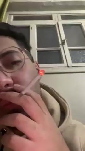 A person is holding a lit cigarette. The person is wearing glasses and a tan hooded sweatshirt.  Smoke is visible as they exhale.  A multi-pane window is in the background.