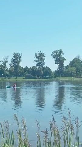 A person in a red hat is in a kayak on a lake, paddling to the left. The lake has tall grass on the right side and trees on the opposite shore. There are three fountains in the lake. The sky is clear and blue.