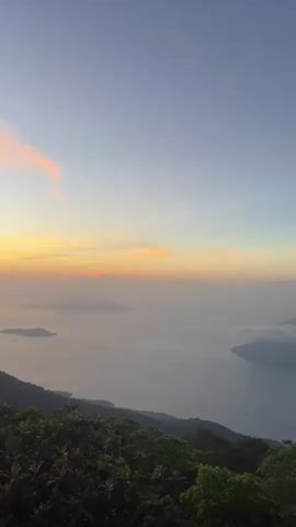 This video shows a panoramic view of a mountainous landscape at sunset. The sky is a gradient of blue, pink, and orange, with wispy clouds. In the foreground, lush green trees cover the slopes of the mountains. In the distance, a body of water is visible, with several islands scattered across it. The sun is setting behind the horizon, casting a warm glow over the scene. The video pans slowly across the landscape, offering a breathtaking view of the natural beauty. The overall mood is serene and tranquil, evoking a sense of peace and wonder.