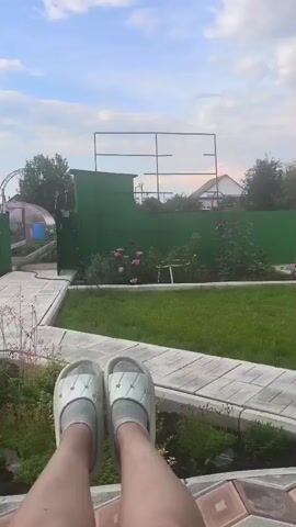 The video shows a first-person perspective of someone sitting outdoors. Their feet, wearing white sliders and gray socks, are visible in the foreground, propped up on a patterned tile surface. Beyond the feet is a well-maintained garden with a green fence, neatly trimmed grass, and various plants. In the background, a house and a greenhouse are visible under a cloudy sky. The camera briefly pans down to show more of the patterned tiles before returning to the garden view.