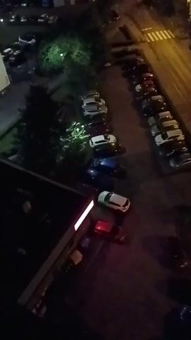 The video shows a nighttime view of a street with parked cars and a crosswalk. The camera is positioned high up, possibly from a window or balcony of a building. In the foreground, there is a portion of a building with a lit window. Several cars are parked in rows on either side of the street. One row of cars is parked along the side of a sidewalk, while another row is parked across the street. The street itself is dimly lit, with streetlights and lights from buildings in the distance creating a yellow glow. The video is shaky and slightly out of focus, suggesting it was taken with a handheld camera.