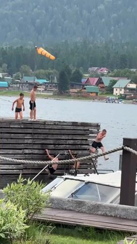 A group of young boys are on a wooden dock over a body of water. Some are standing on top of the dock, while others are climbing down the side. A boat is docked below the pier. In the background, there are houses and trees on a hillside. An orange windsock is visible on a pole.