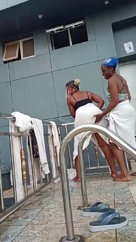 Here's a description of the video:

The video shows two women by a swimming pool. Both are partially covered in white towels. One woman has her back to the camera, wearing a black top and a towel wrapped around her waist. The other woman is standing behind her, smiling and with her hands on the first woman's buttocks. She wears a striped top and a blue head wrap. They are on a tiled deck next to the pool, with a metal railing separating them from the pool area. There are also several towels hanging over the railing. The background features a grey building with windows. Upbeat music plays in the background.