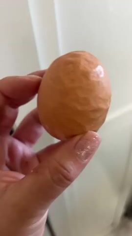 The video shows a close-up of a person's hand holding an egg. The egg is light brown and appears to have a slightly textured or wrinkled surface, possibly due to being boiled or otherwise cooked. There are also some shiny patches on the egg's surface, which could be water droplets or oil. The hand holding the egg is positioned in the foreground, with the fingers and thumb wrapped around the egg. The background is blurred and appears to be an indoor setting, with a white door or wall visible. The lighting suggests it is daytime. The overall impression is a simple, everyday action of holding an egg. There is no visible text in the video.